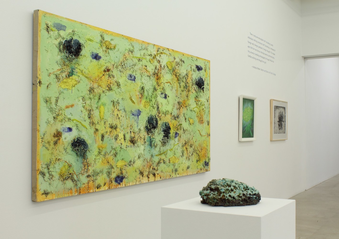 Photograph of a detail view of an exhibition, with one large painting and two smaller paintings hung on a white wall, with an organic-looking sculpture on a pedestal in front