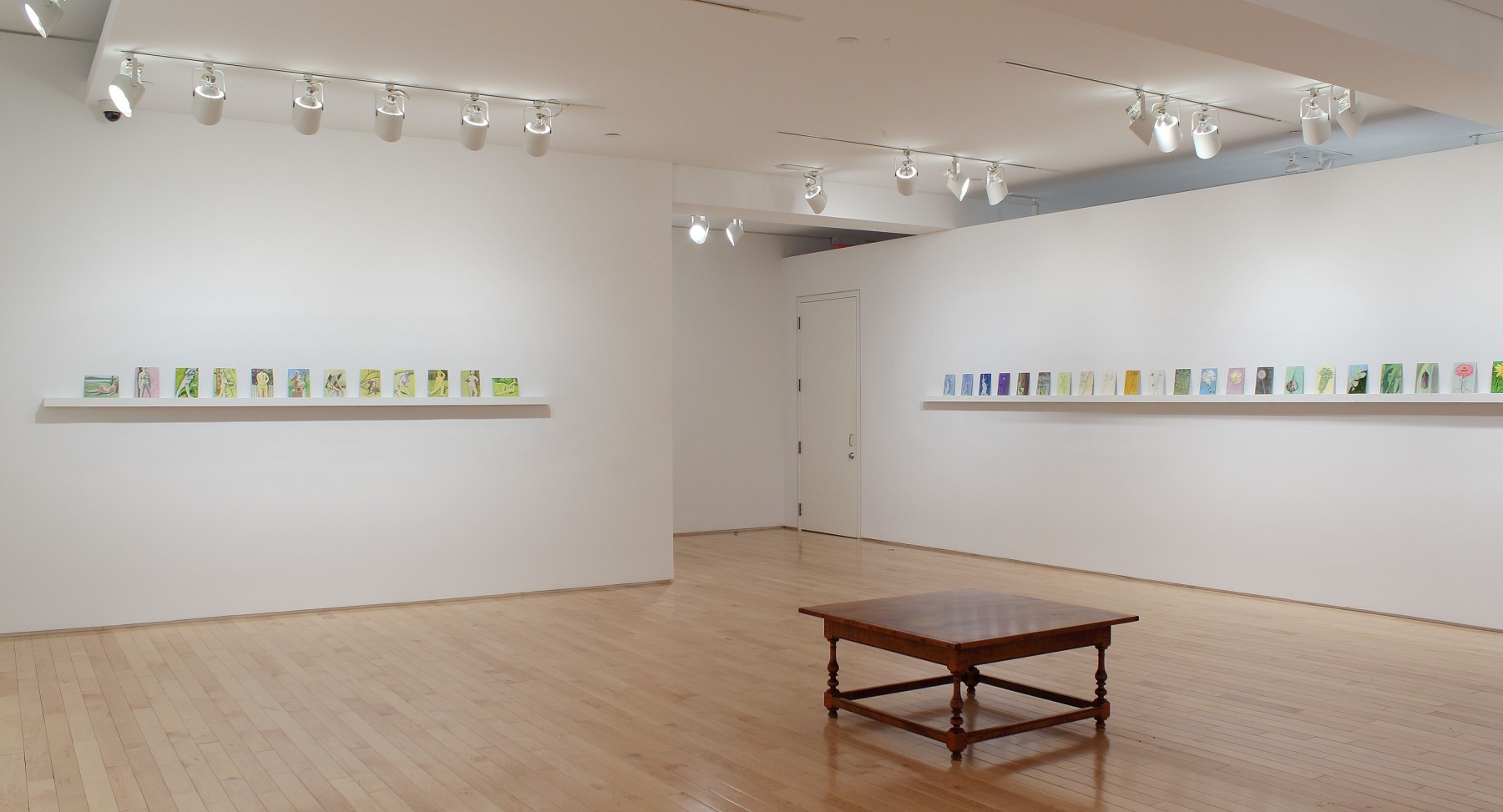Photograph showing a corner view of an exhibition space, each wall holding dozens of small paintings on a long shelf