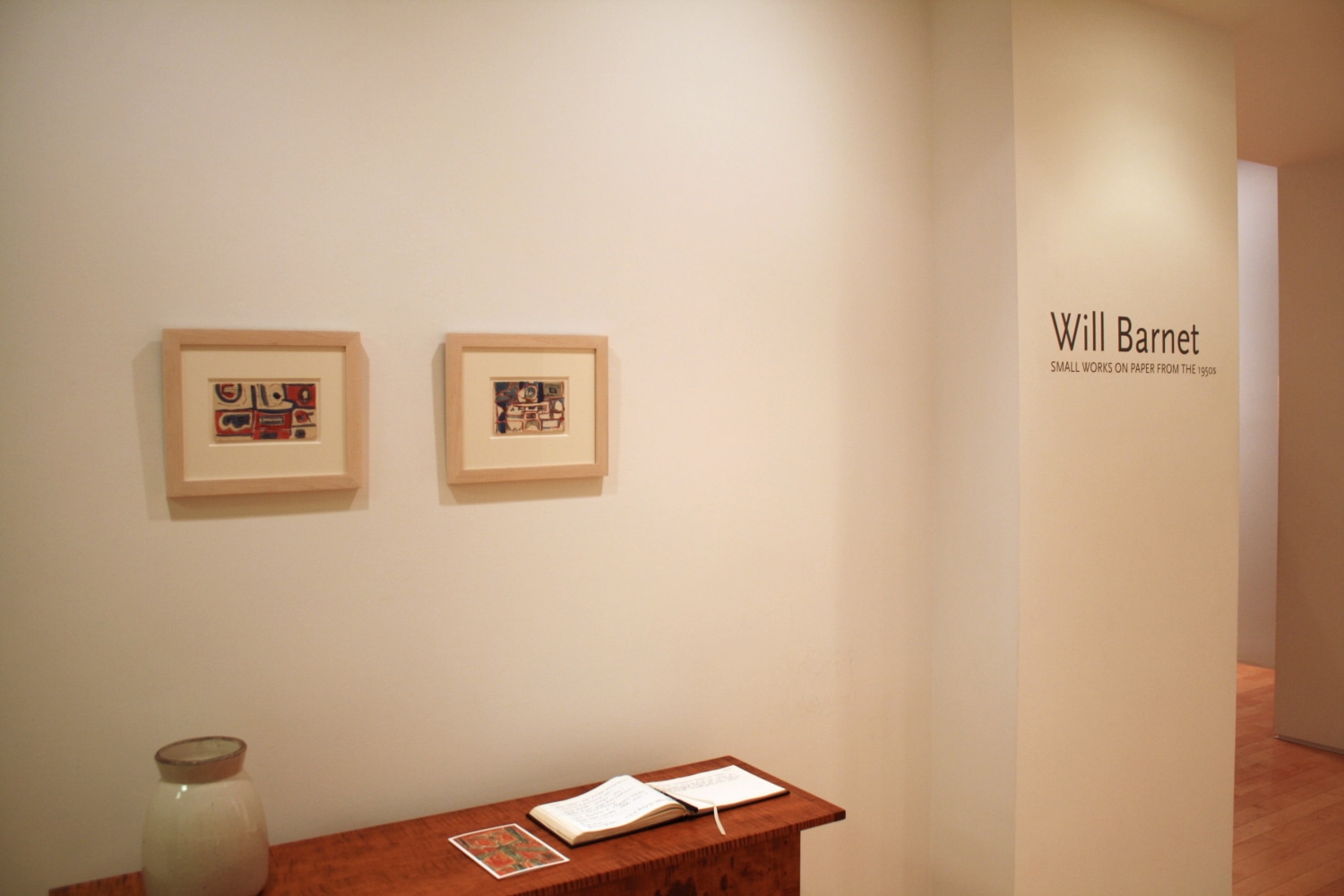 Photograph showing a view of an exhibition space, with two small framed paintings hanging over a wooden table, and wall text that reads &quot;Will Barnet: Small works on paper from the 1950s&quot; on the wall to the right