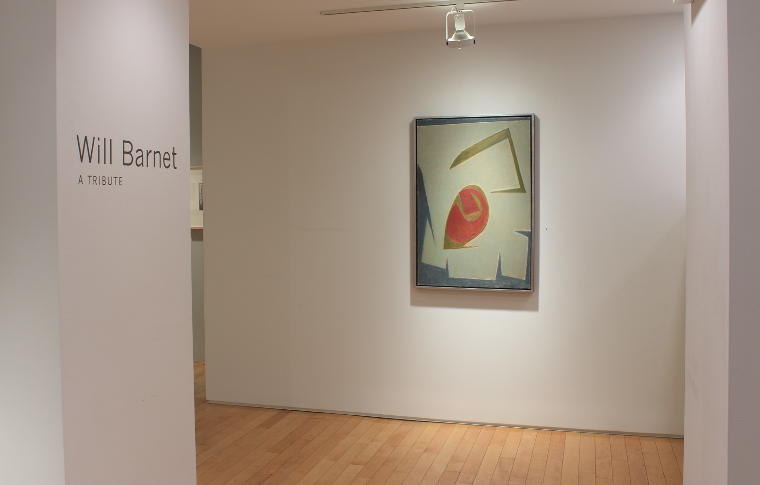 Photograph showing a view of an exhibition space with one painting hanging on the wall and wall text that reads &quot;Will Barnet: A Tribute&quot; on the left