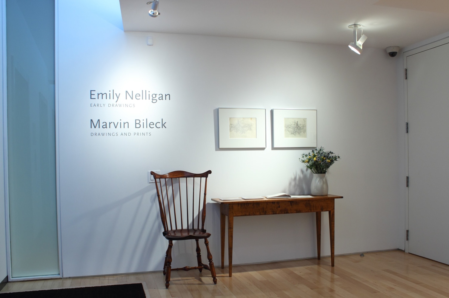 Photograph showing a view of an exhibition space, with two framed drawings hanging on a white wall above a small table and chair