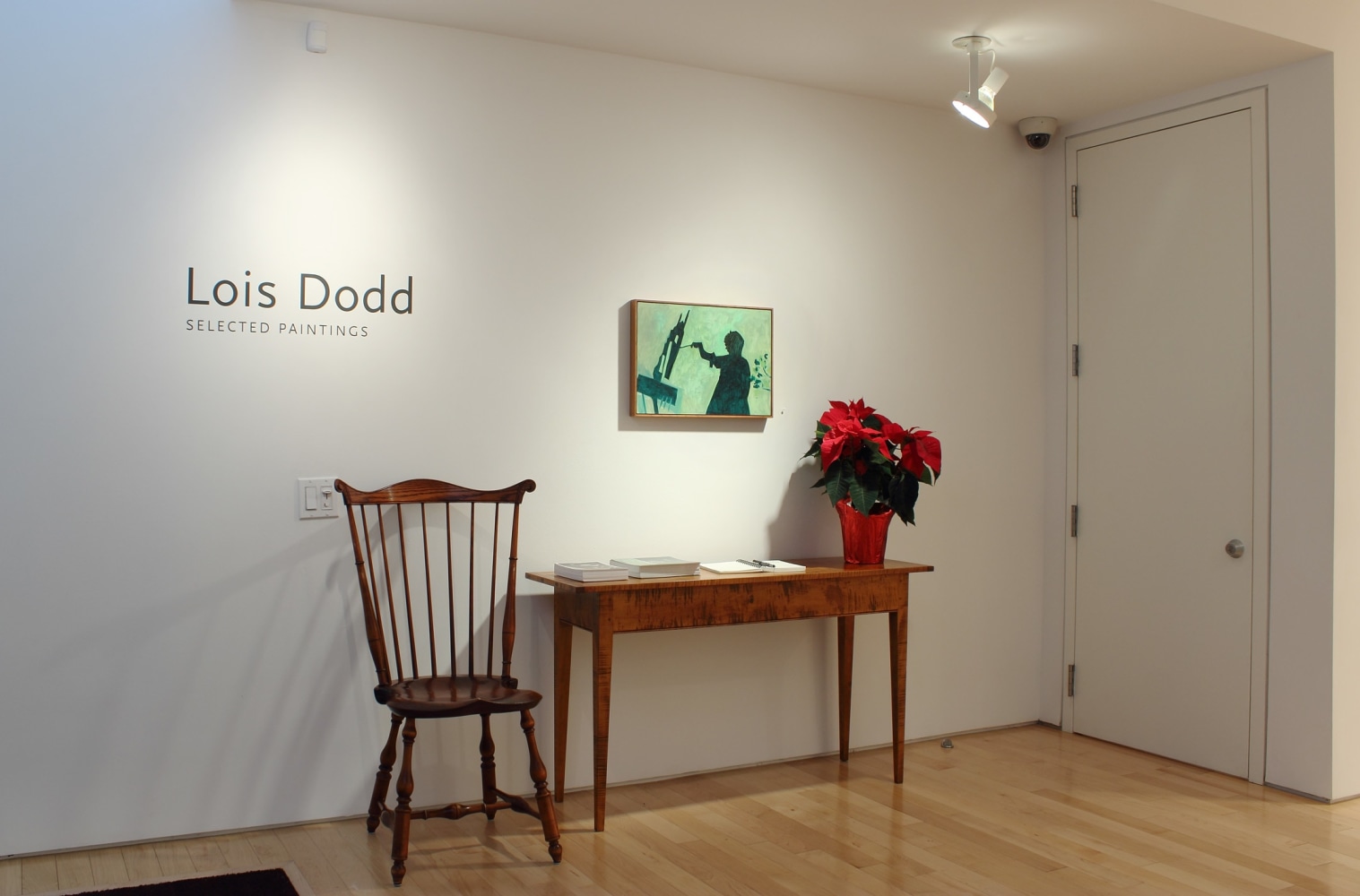 Photograph showing a view of an exhibition space, with a painting hanging on a white wall above a table and chair
