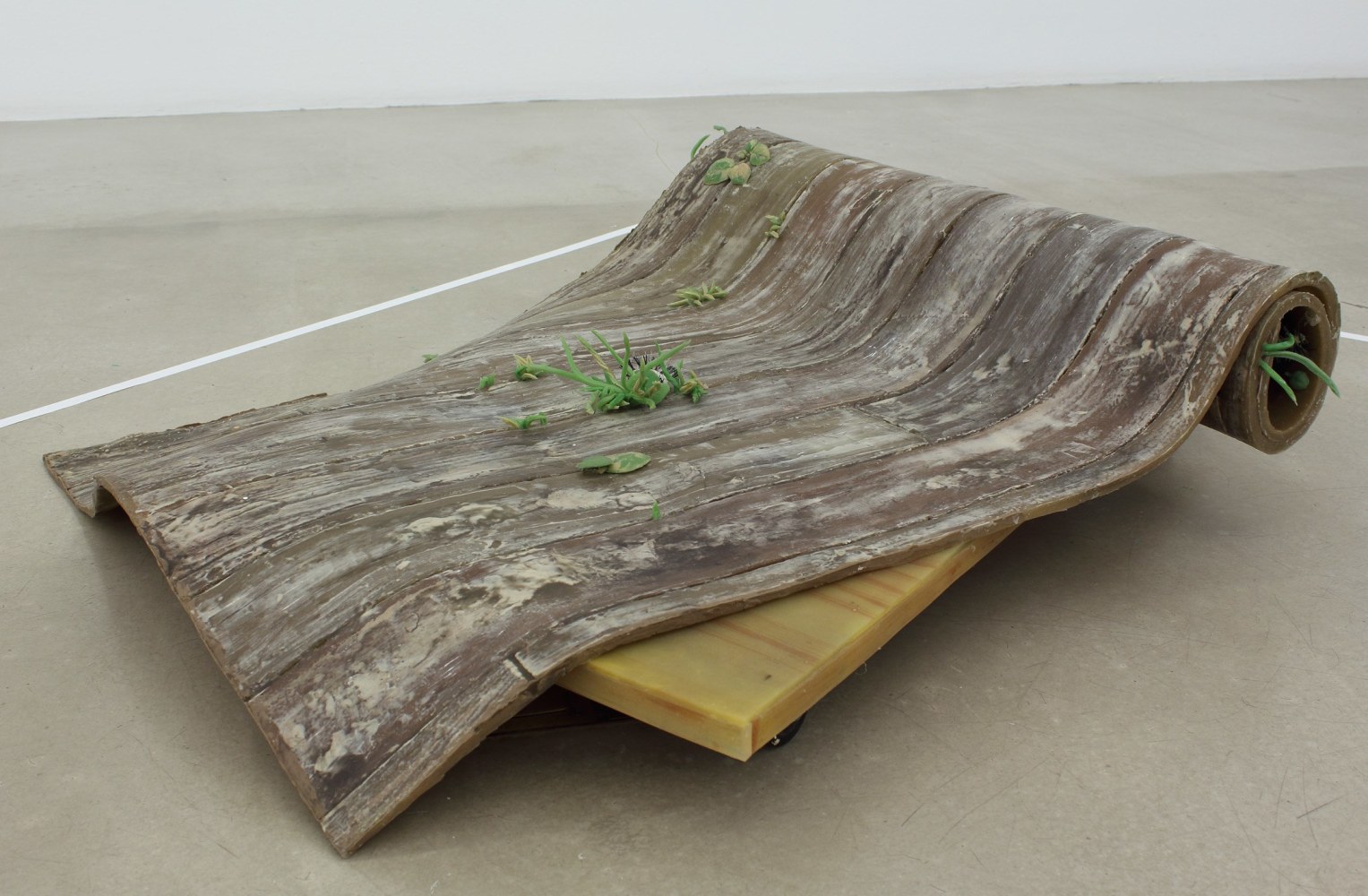 Photograph of a realistic rubber sculpture of a wooden-looking floor mat with weeds growing out of the cracks, rolled up and sitting on top of a dolly with wheels