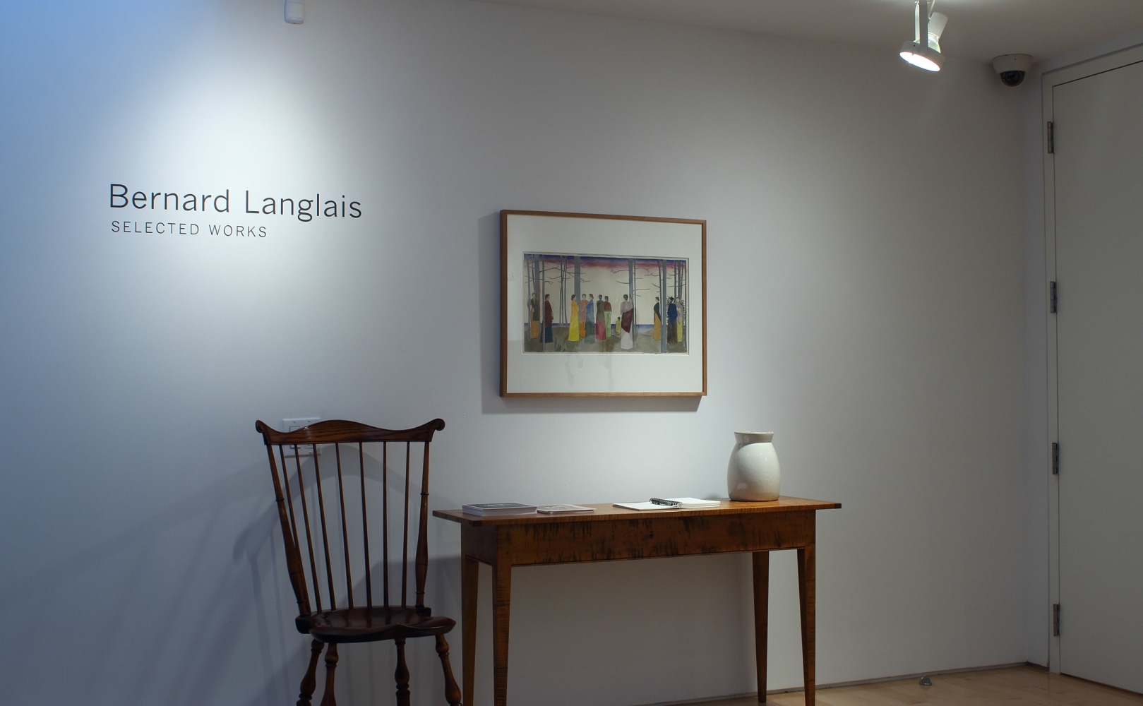 Photograph showing a view of an exhibition space, with a framed drawing hangin on a white wall over a table and chair