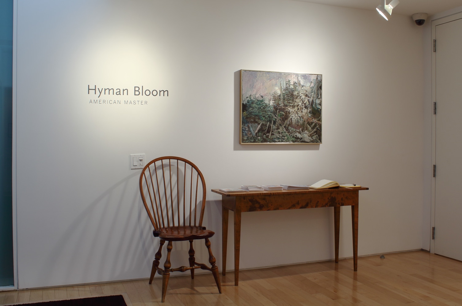 Photograph showing a front view of an exhibition space with one painting hanging on a white wall above a chair and side table