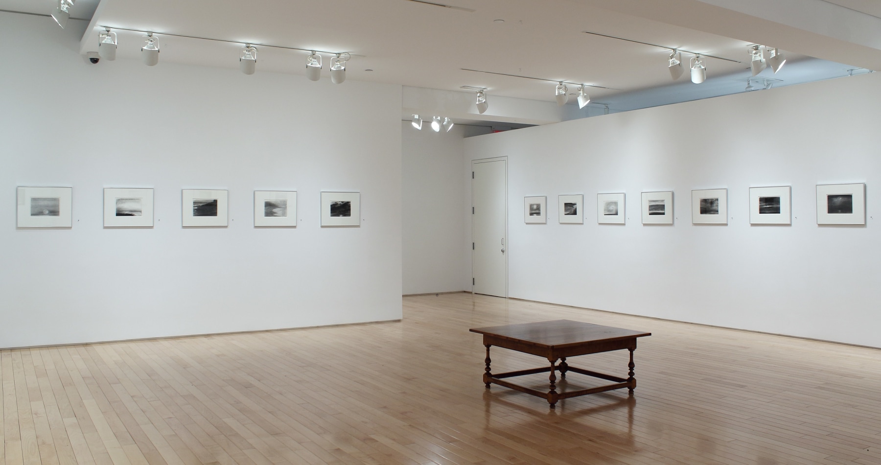 Photograph showing a corner view of an exhibition space, with twelve framed drawings hanging on white walls