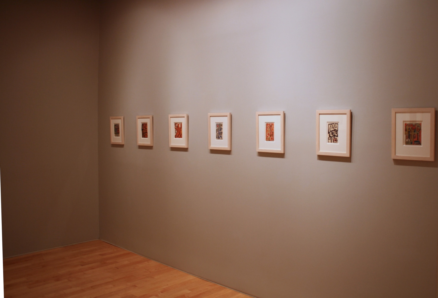 Photograph showing a corner view of an exhibition space, with seven framed paintings hanging on a grey wall