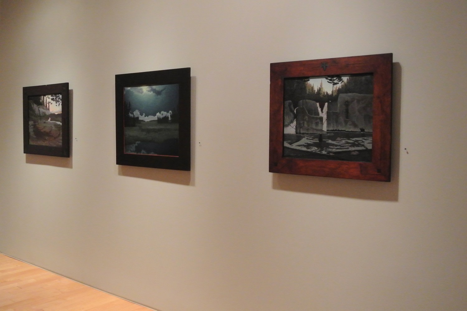 Photograph showing a view of an exhibition space, with three framed paintings hanging on a grey wall