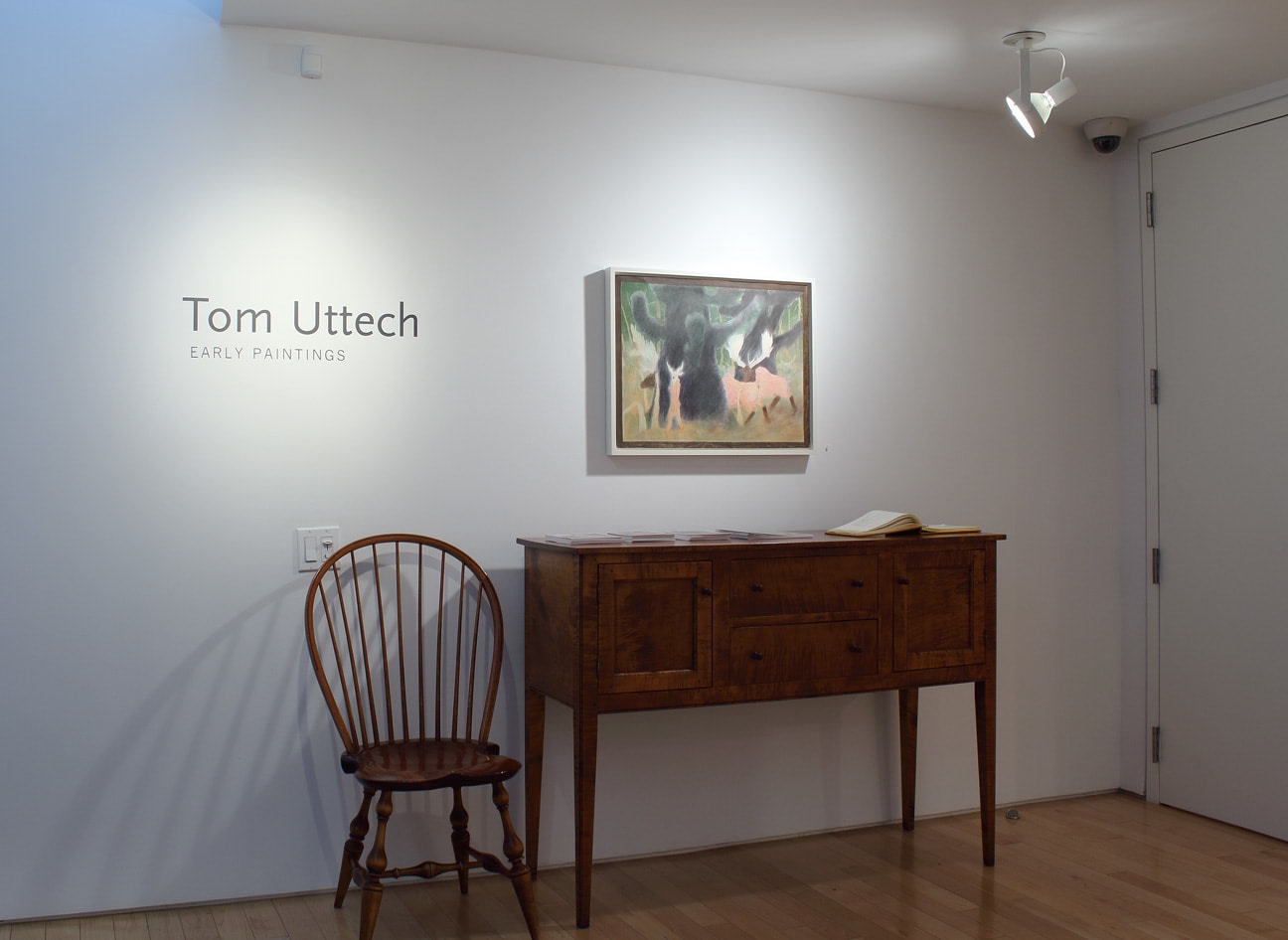 Photograph showing a front view of an exhibition space with a drawing hanging on a white wall over a side table and chair