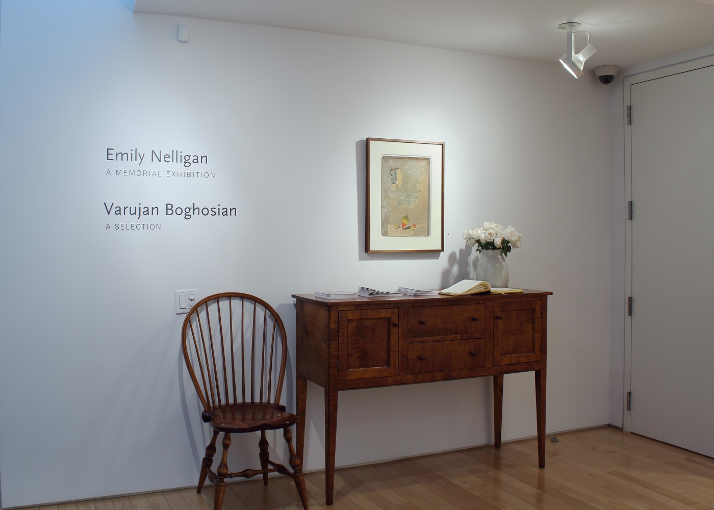 Photograph showing a front view of an exhibition space, with one painting hanging on a white wall above a table and chair
