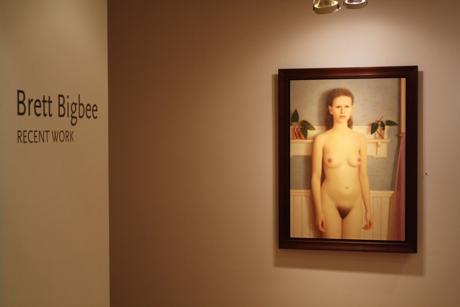 Photograph showing a view of an exhibition, with one framed painting hanging on the wall to the right, and wall text that reads &quot;Brett Bigbee: Recent Work&quot; on the wall to the left