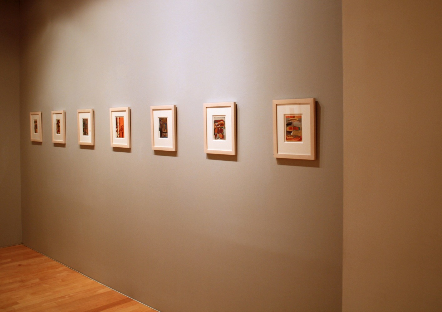 Photograph showing a view of an exhibition space, with seven small framed painting hanging on a grey wall