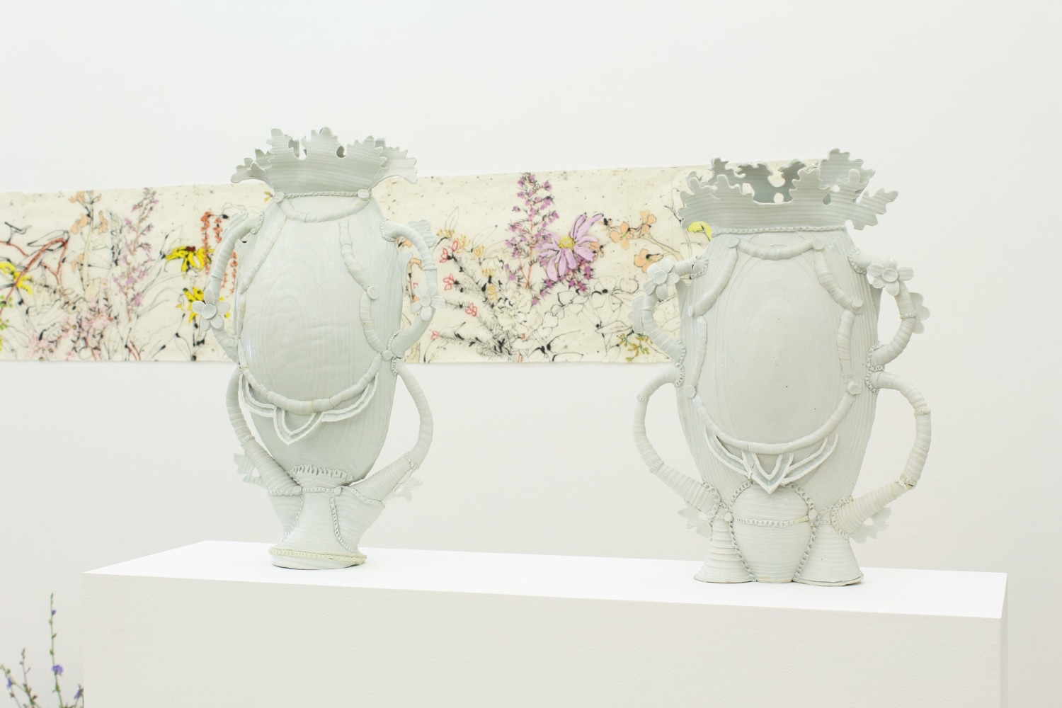 Photograph showing a detail of an exhibition, with two white porcelain sculptures in the foreground and a drawing on the wall in the background.