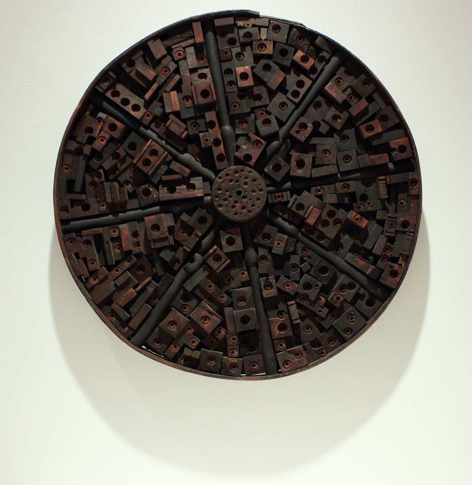 A circular, wall-hanging sculpture composed of pieces of wood and metal assembled together