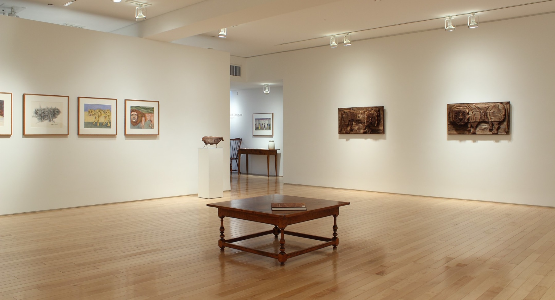 Photograph showing a corner view of an exhibition space, with three framed drawings and two wooden sculptures hanging on white walls