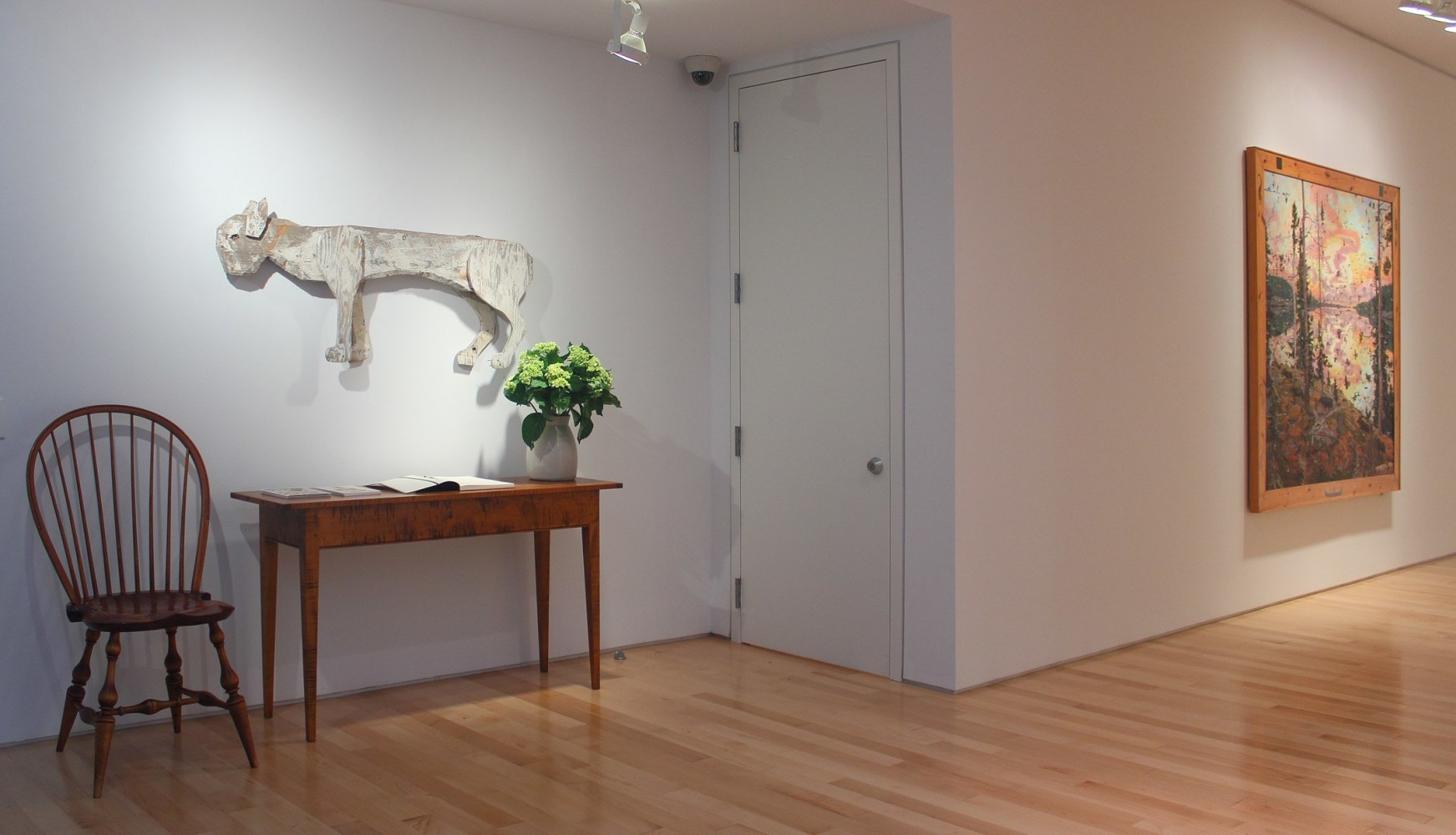 Photograph showing a view of an exhibition space with a wooden sculpture hanging over a table and chair