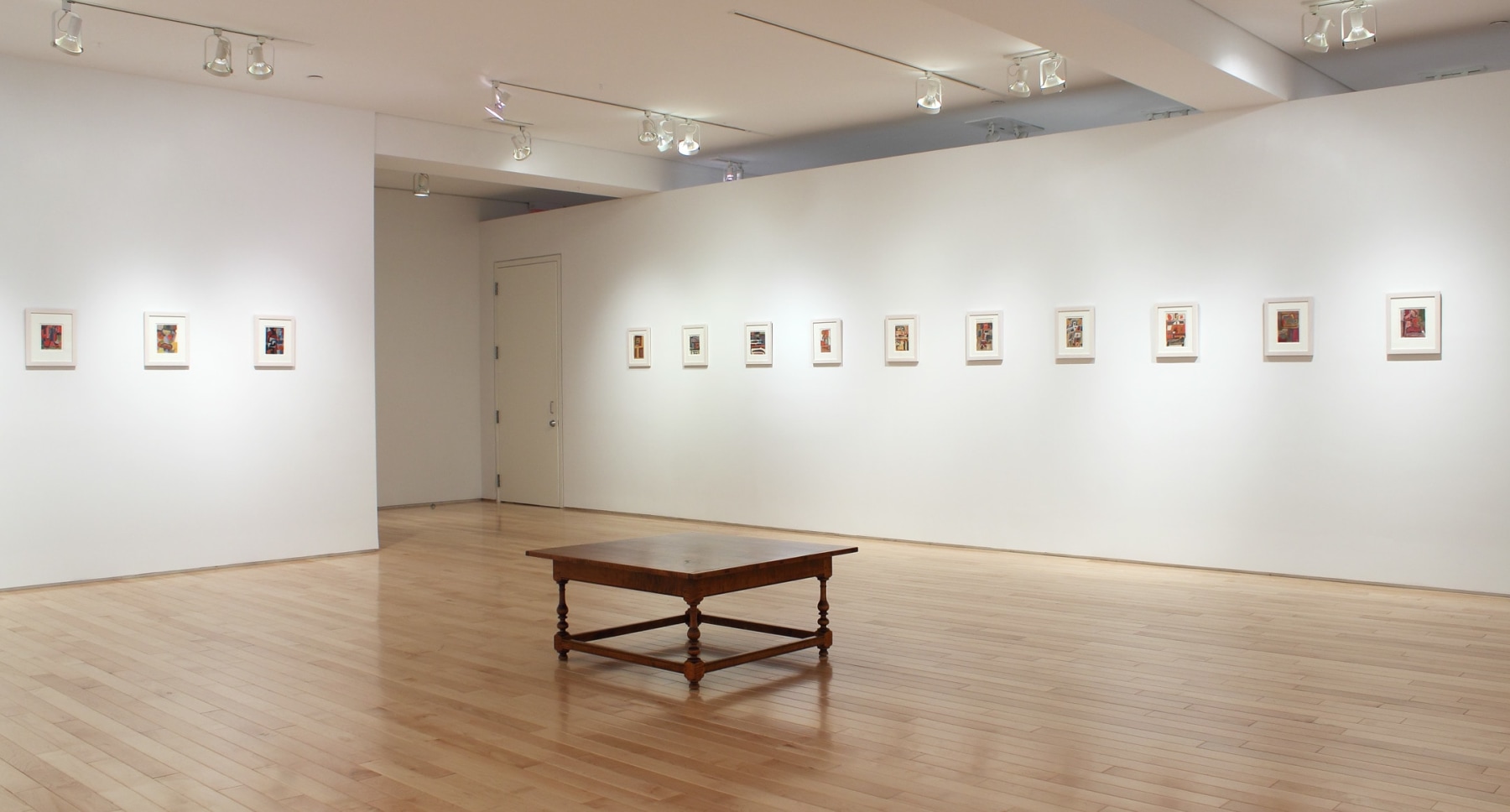 Photograph showing a corner view of an exhibition space, with thirteen small framed paintings hanging on white walls