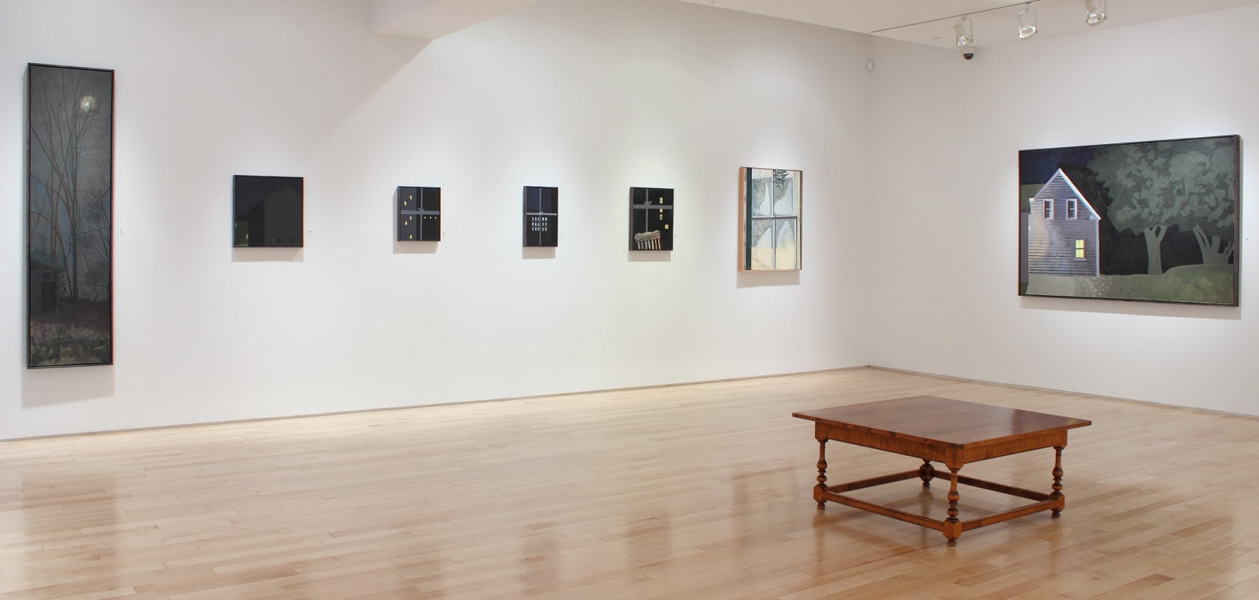 Photograph showing a view of an exhibition space with seven paintings hanging on a white wall