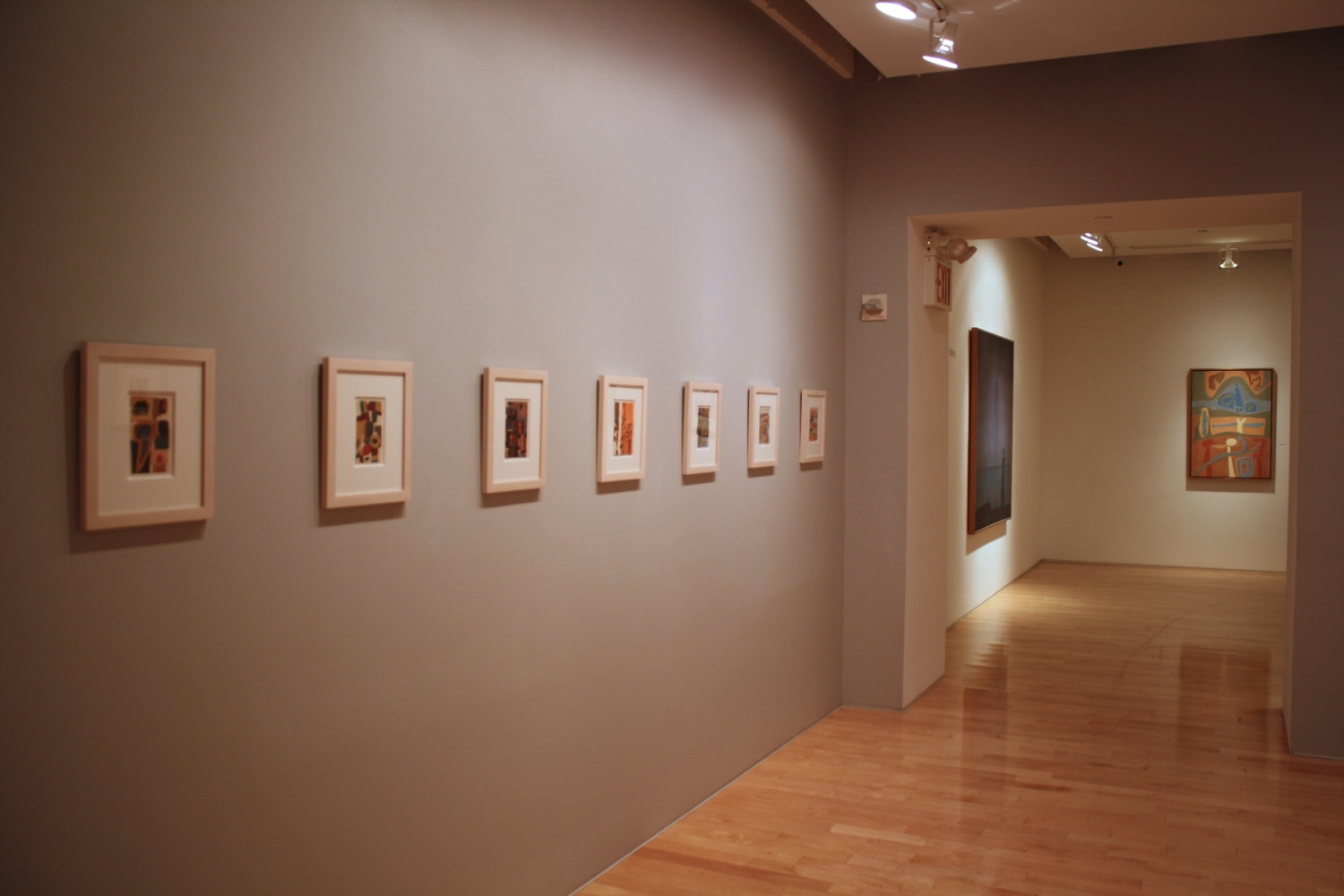 Photograph showing a corner view of an exhibition, with seven small framed paintings hanging on a grey wall