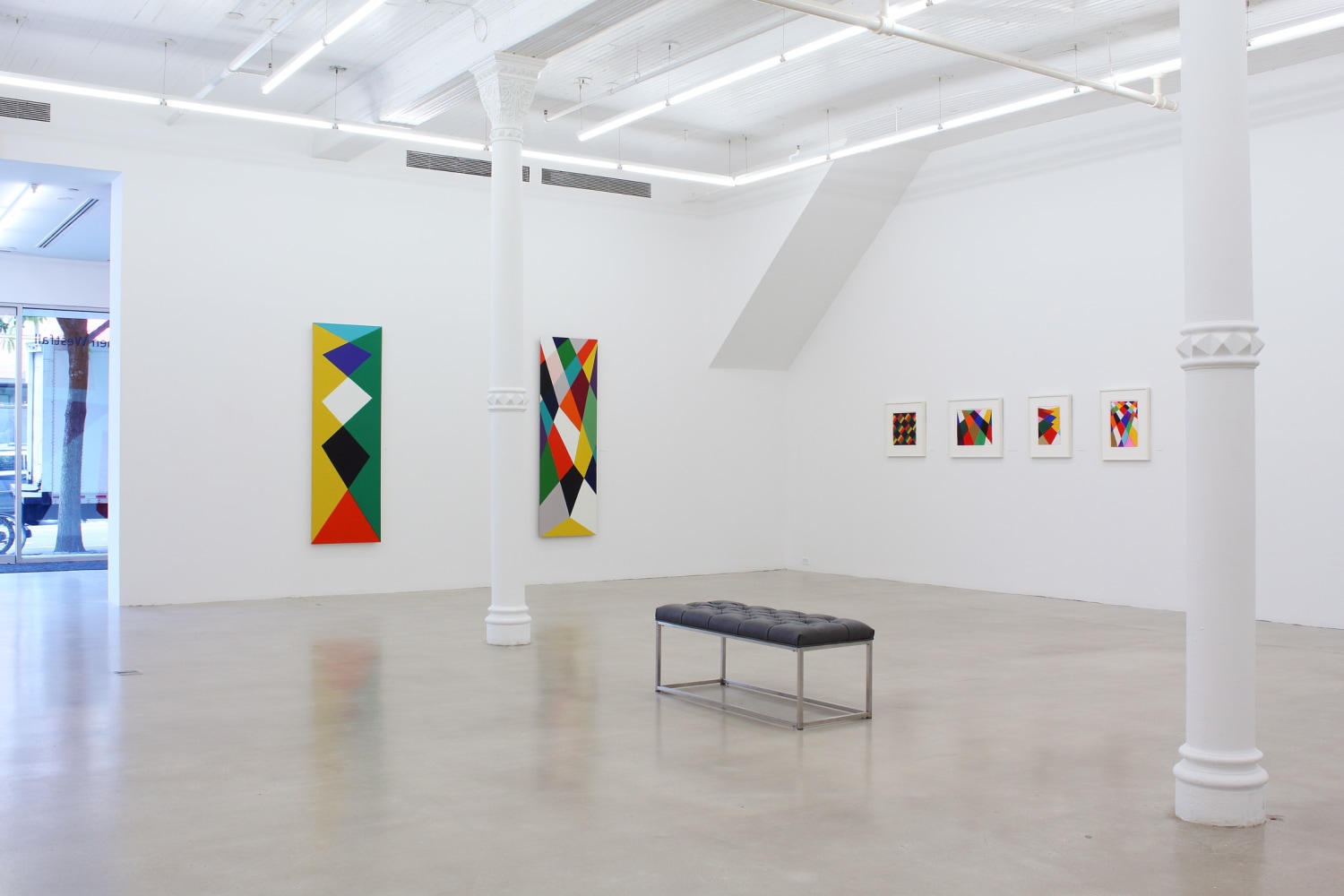Photograph showing a corner view of an exhibition space, with six paintings hanging on white walls