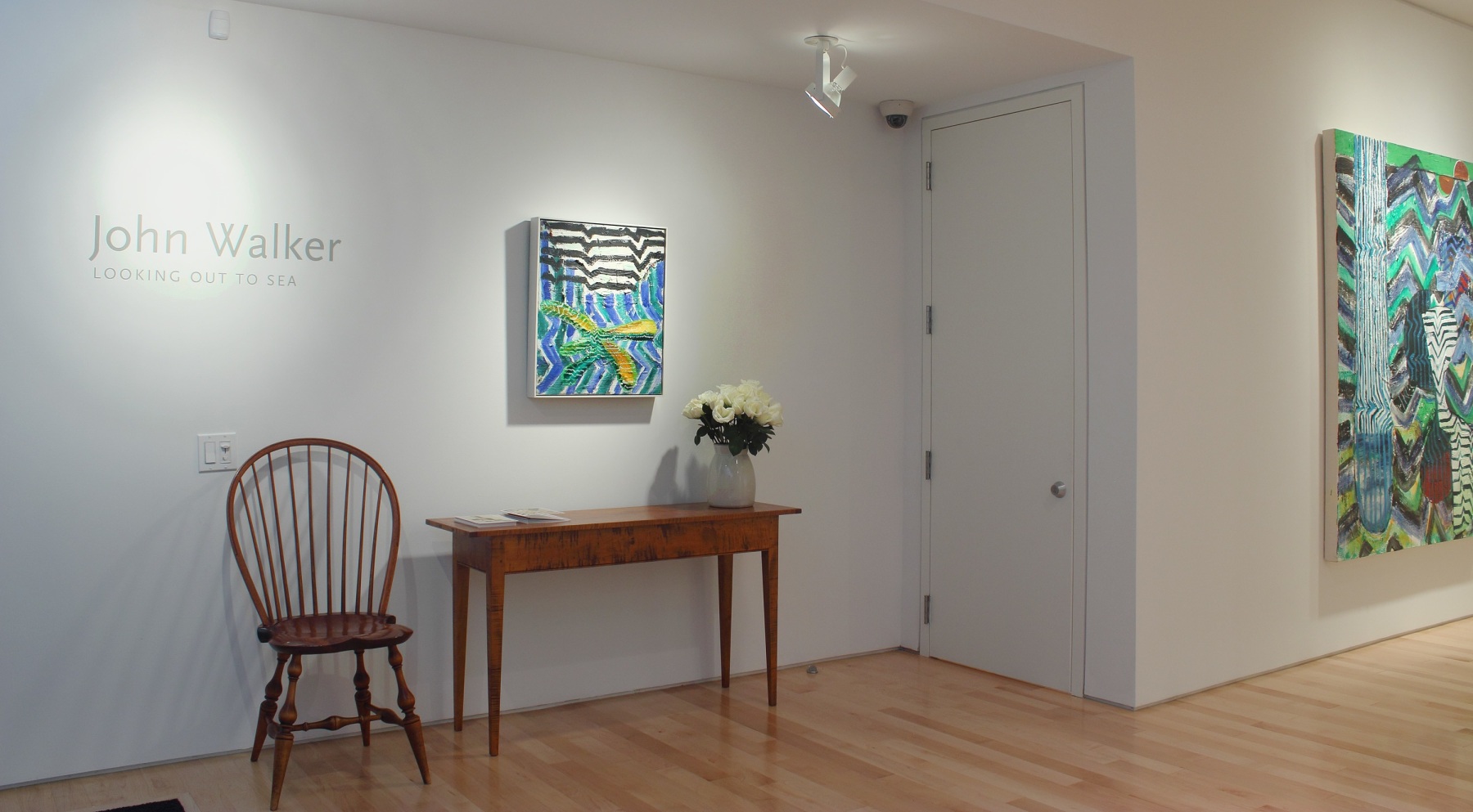 Photograph showing a view of an exhibition space, with one small painting hanging on a white wall over a table and chair and wall text reading &quot;John Walker: Looking out to Sea&quot; to the left