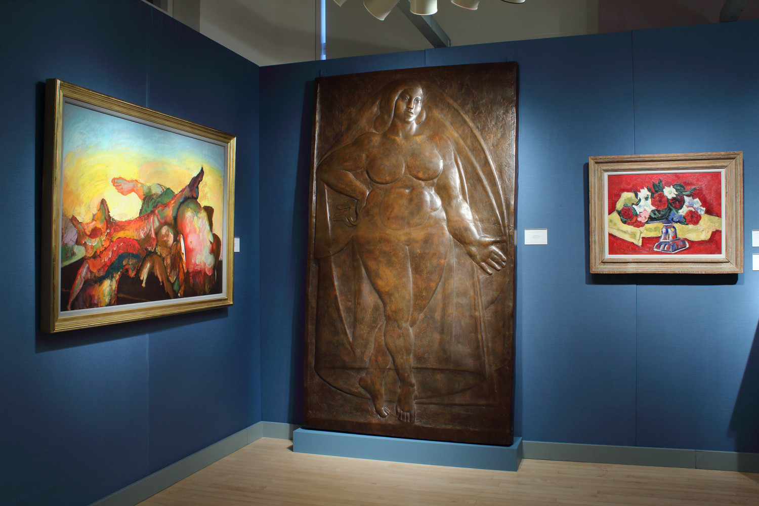 Photograph showing a corner view of an exhibition booth, with one relief sculpture standing and two paintings hung on blue walls