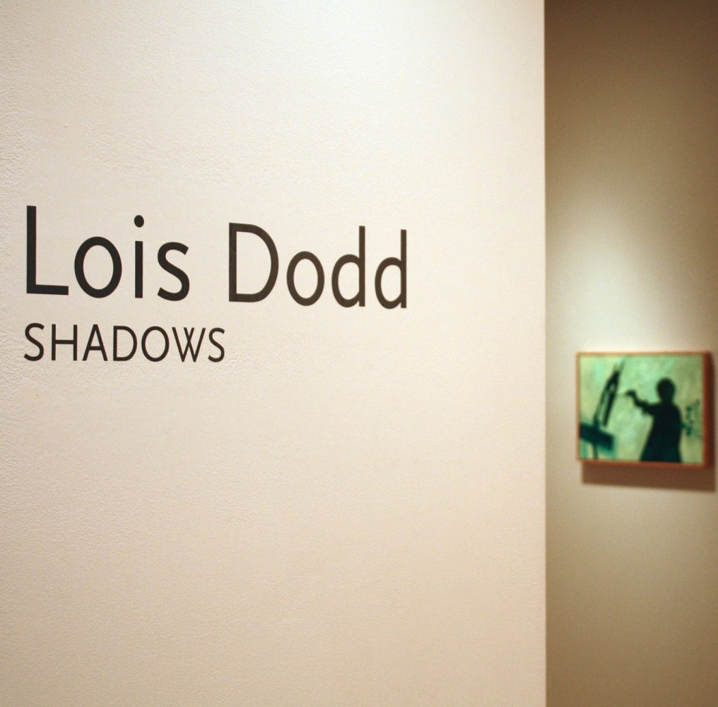Photograph showing a view of an exhibition space, with wall text that reads &quot;Lois Dodd: Shadows&quot; on the wall to the left, and a small painting hanging on a wall in the background