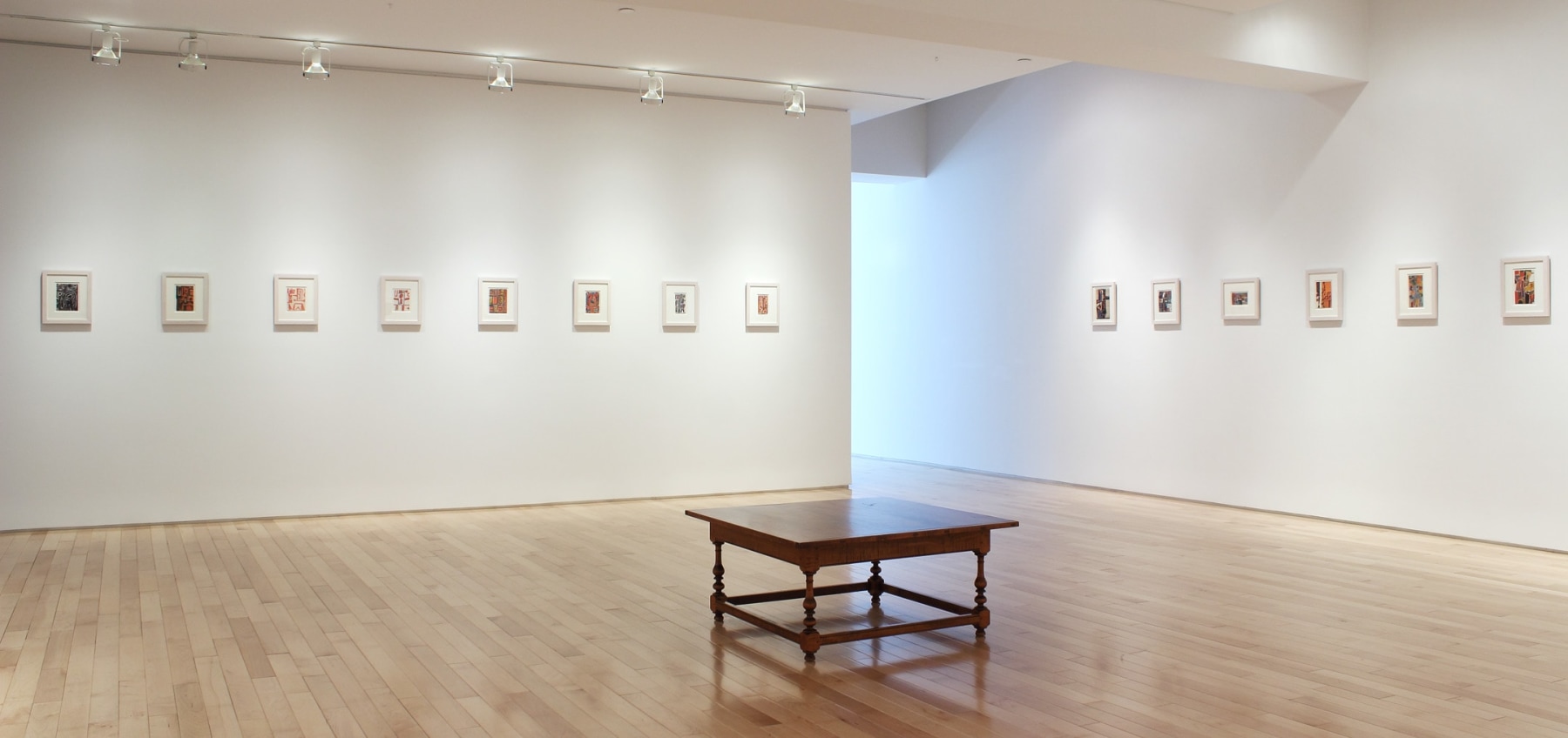 Photograph showing a corner view of an exhibition space, with fourteen small framed paintings hanging on white walls