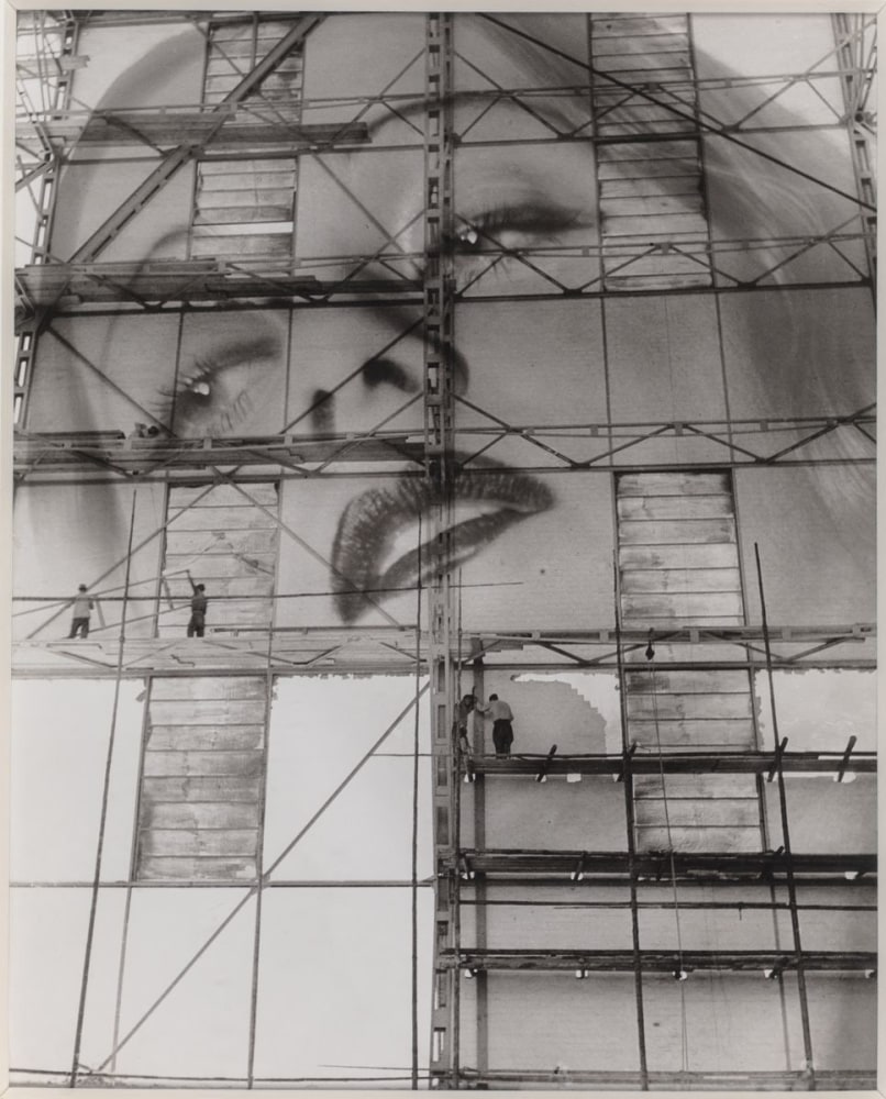 A model's face is painted onto the facade of a building by painters working on scaffolding in this black and white photo.