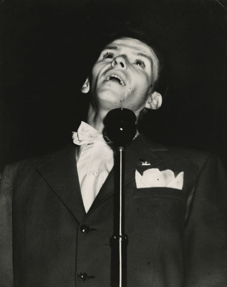 Black and white photographic print of Frank Sinatra singing behind a microphone at the Palace Theatre by Weegee