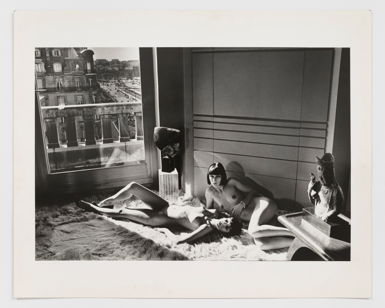 Black and white photographic by Helmut Newton featuring a nude woman crouched beside a bed with a chain around her neck in a hotel room