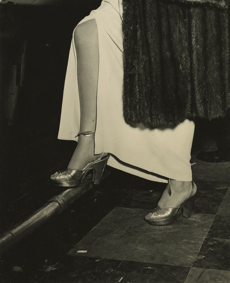 A black and white photographic print by Weegee of the lower half of a woman at a bar wearing heels, a dress with a slit, and fur coat with one foot lifted on the foot rail
