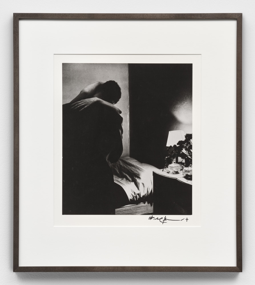 A black and white photographic print by Bill Brandt depicting the back of a figure with the arms of another person wrapped around their upper body in a bedroom