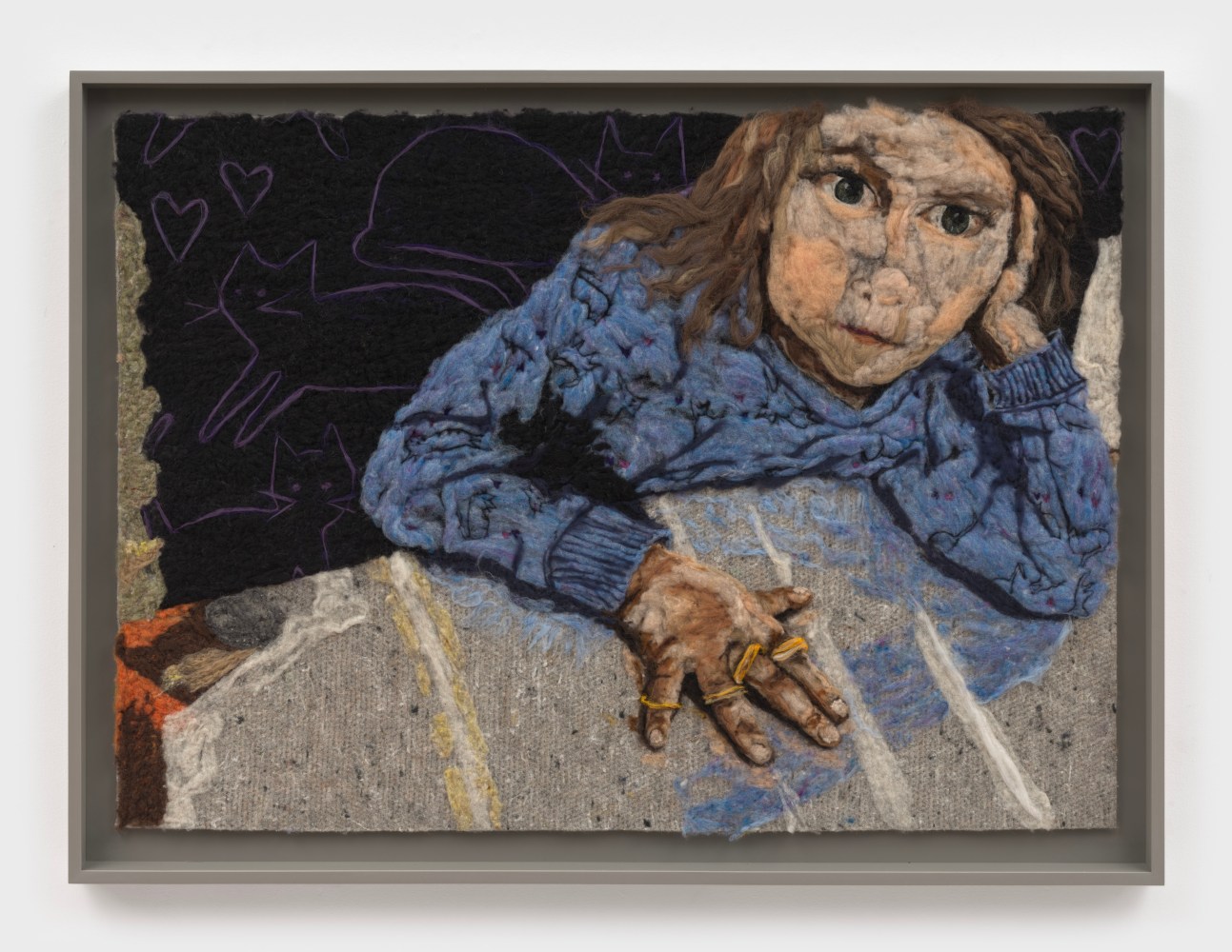 A felted wool portrait of a young girl in a blue sweater leaning on a table wearing four golden rings.