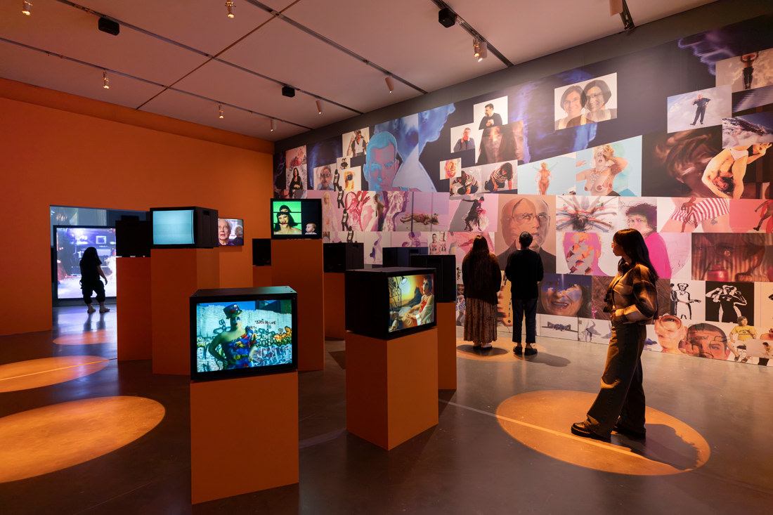 Museum exhibition space with multiple video monitors and a wall filled with various stills of video portraits