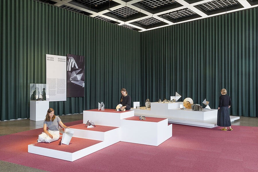Museum installation of sculptures on top of white plinths with pink carpeting and green curtains