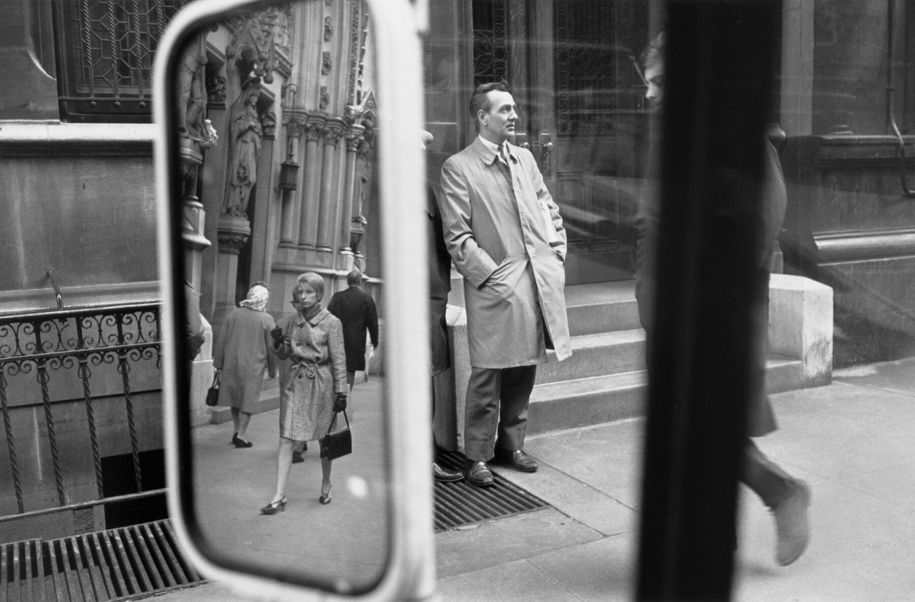 black and white photo of a street taken from a car with the rear view mirror reflecting the other side of the sidewalk, 5 pedestrians