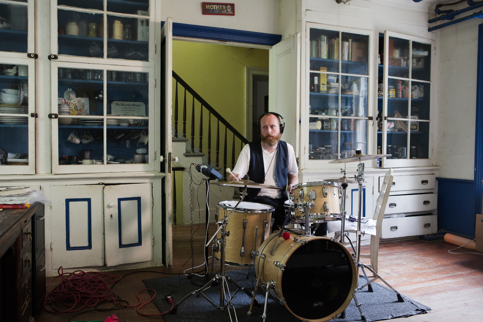 A bearded man playing drums in front of white cupboards with blue trim