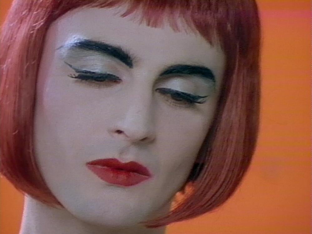 Close-up video still of a performer in a burgundy bobbed wig