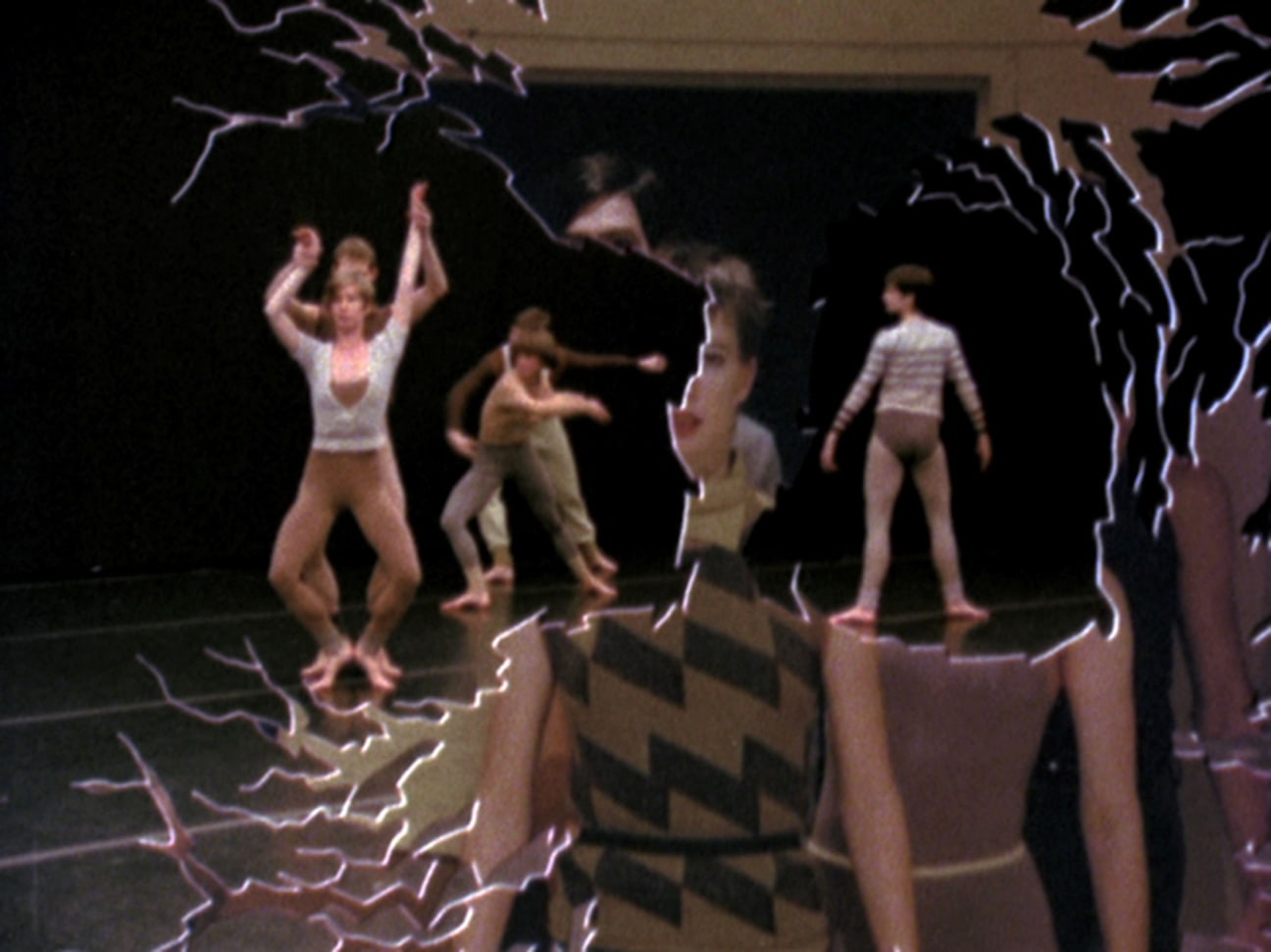 Video still of a dance performance. Dancers are wearing various shades of tan and are against a black background