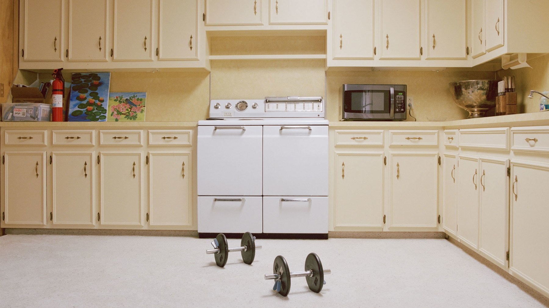 An empty kitchen with yellow cabinets and 2 barbells on the floor in front of a stove