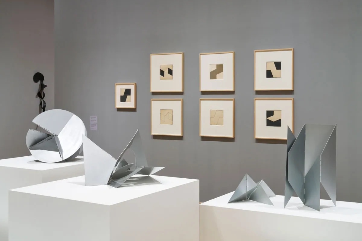 A Lygia Clark installation: 4 metal sculptures on plinths with 7 black and white collages hung on a grey wall in the background