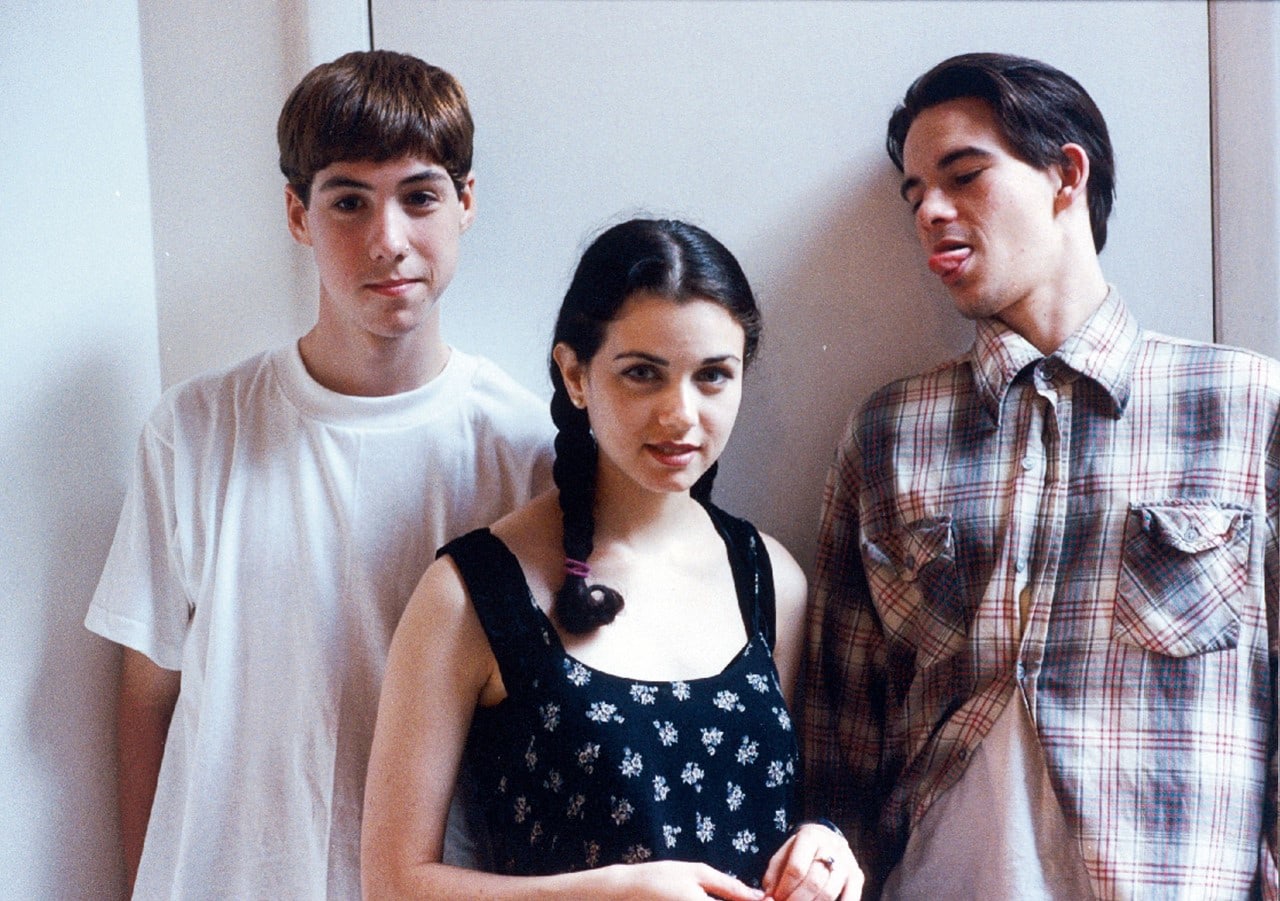 3 teenagers standing against a white wall: 2 boys and 1 girl, the boy on the right side stocks his tongue out at the girl