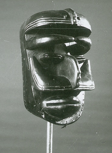 Black and white photo of a mask replicating a human profile, made of wood, brass tacks, black pigment, string,  and 2 nails. The facial features of the profile are carved into the mask so that the scalp, prominent eyebrows, eyes, hooked nose, cheekbones, and lips all protrude from the base of the mask.