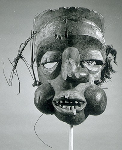 Mask made of wood, stain, reed, and fabric, replicating a human profile, with eyes carved out beneath the eyelids. The mouth is open, bearing teeth, and its bulbous cheeks and twisted nose greatly protrude from the mask.