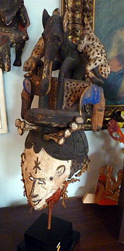 Polychrome wooden mask replicating the human profile. The features of the face are outlined in brown, including a six point star in the center of the forehead. Resting on top of the figure's hair is a wooden black horse, flanked on both sides by leopards that climb down its side. Resting in between the legs of the horse is a spotted, double-faced antelope.