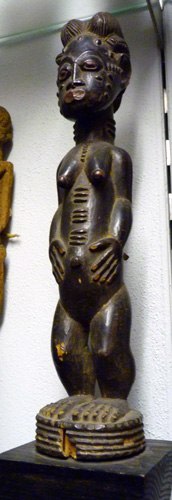 Wooden sculpture of the elongated and curved body of a nude woman, standing and holding her belly. Her body, especially her torso exhibits scarification nodes and she wears a bulbous, lobed coiffure.