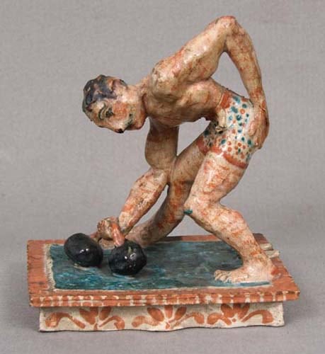Sculpture of a man bending over to lift a small weight standing on a teal mat. His skin is painted a splotchy red and his hair is black. He is wearing blue and red polka dot shorts and as he bends over one hand rests on the back of his shorts. The weights have one black ball on each end of a white rope. The sculpture is mounted on a red and white decorative block.