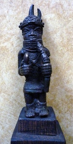Bronze sculpture of a figure seated on a stool, situated on a wooden base. He is dressed and holding a club (viewer's left) and staff (viewer's right) in either hand. He is adorned with ringed jewelry around his neck and ankles, and chest. Above his enlarged eyes is his knitted headdress, complete with horns.