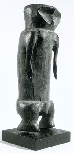Abstract wooden statue of a standing male complete with an exaggeratedly cylindrical torso, short arms, and wide small legs. No prominent physical features are depicted.